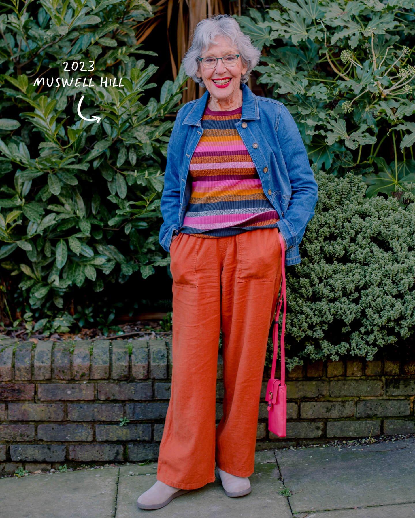 an older woman standing on a sidewalk in front of a bush wearing White Stuff
