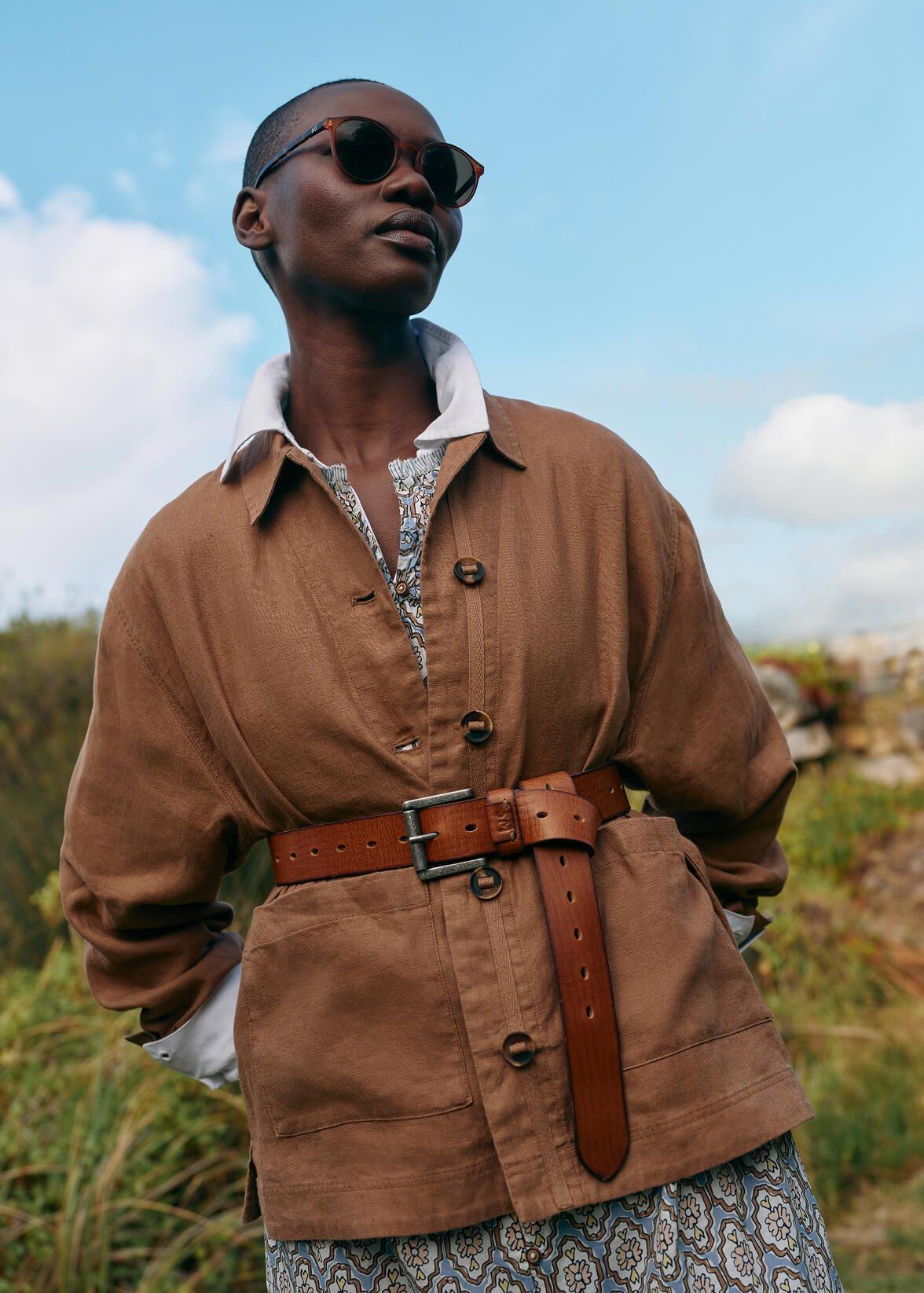 a woman in a brown jacket and sunglasses standing in a field