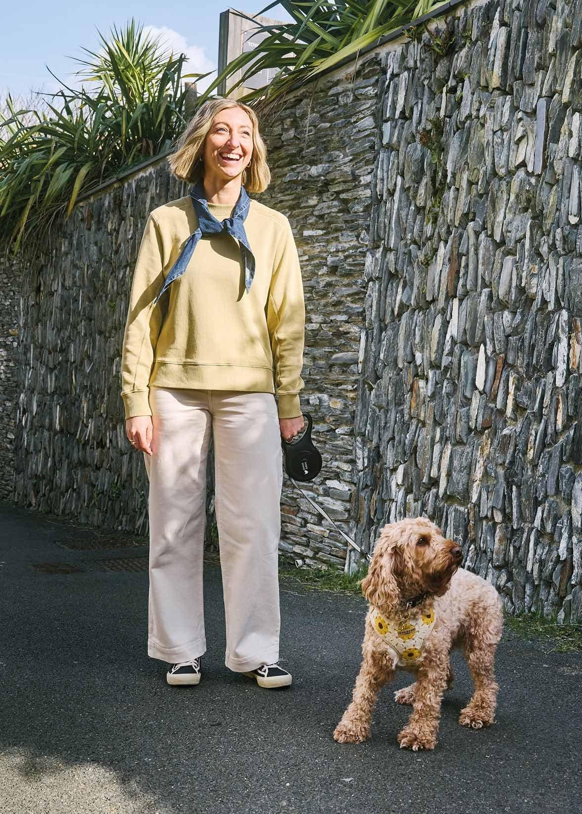 a woman in sweater with a small dog on a leash in front of a stone wall