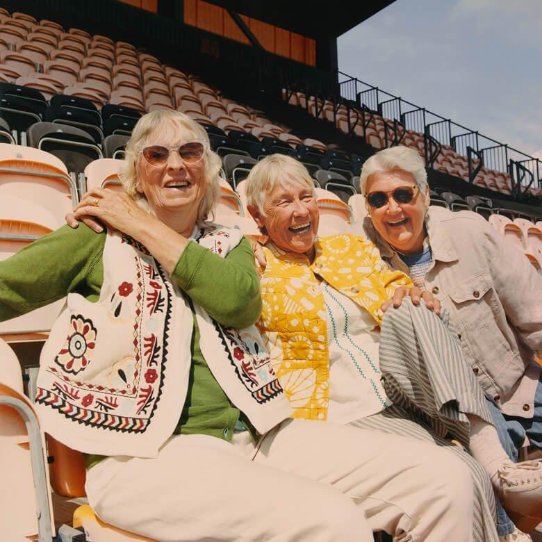 three older women sitting on bleachers wearing White Stuff clothing