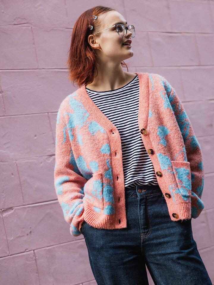 a woman in a striped shirt and cardigan standing against a pink wall