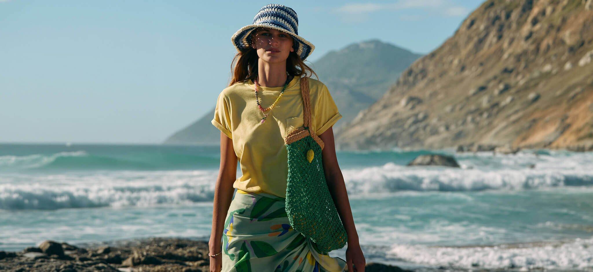 A woman stood a beach wearing a floral sarong, yellow t-shirt and a green woven tote bag and blue and white bucket hat