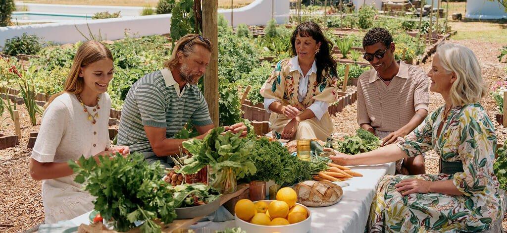 A group of people sat around a table outside wearing clothes from the White Stuff summer collection. On the table is an array of fruit and vegetables.