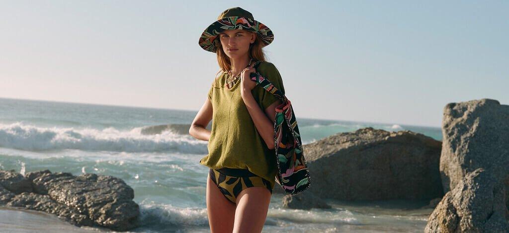 A woman stood on a beach wearing a green tshirt and a leafy printed pair of bikini bottoms styled with a matching embroidered tote bag and bucket hat