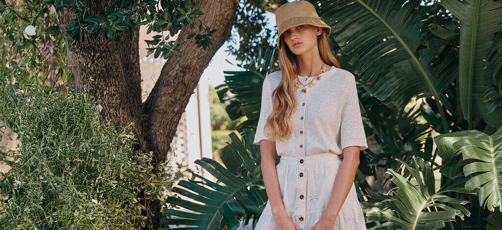 A woman stood outside with a cream button up top and a midi skirt styled with light brown bucket hat and gold chunky necklace