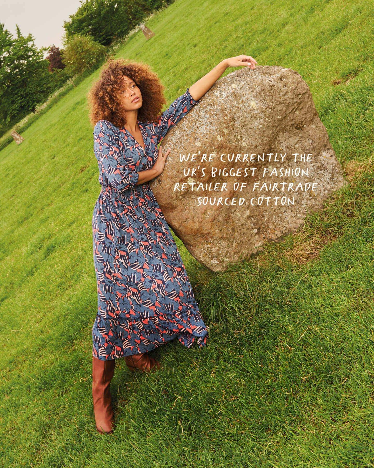 Woman leaning over rock in blue and pink patterned dress and brown heeled boots.