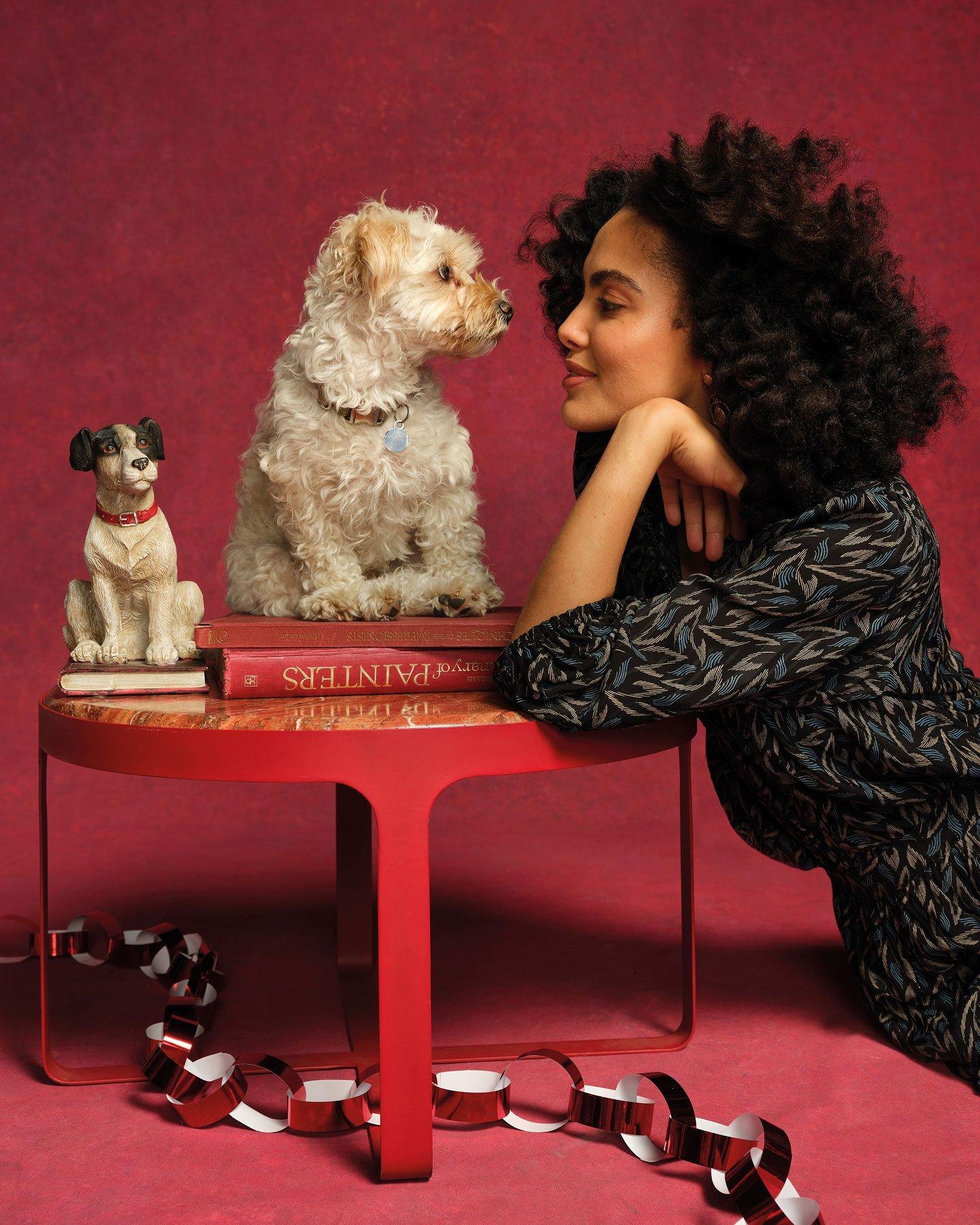 a woman leaning on a round red table looking into the eyes of a dog sat on a couple of books next to a statue of a dog on a book. A decorative paperchain is on the floor