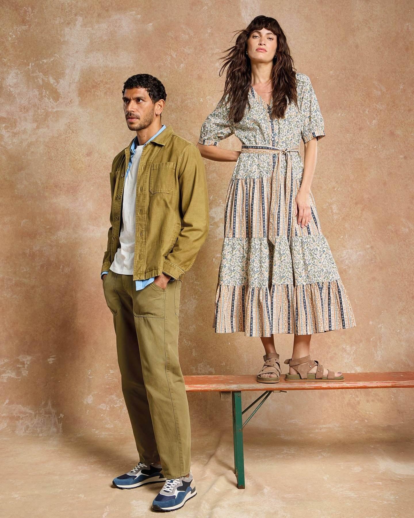 A female and male model standing next to each other. Female model wearing patterned midi dress and male model wearing khaki chinos, white t-shirt & khaki button up jacket.