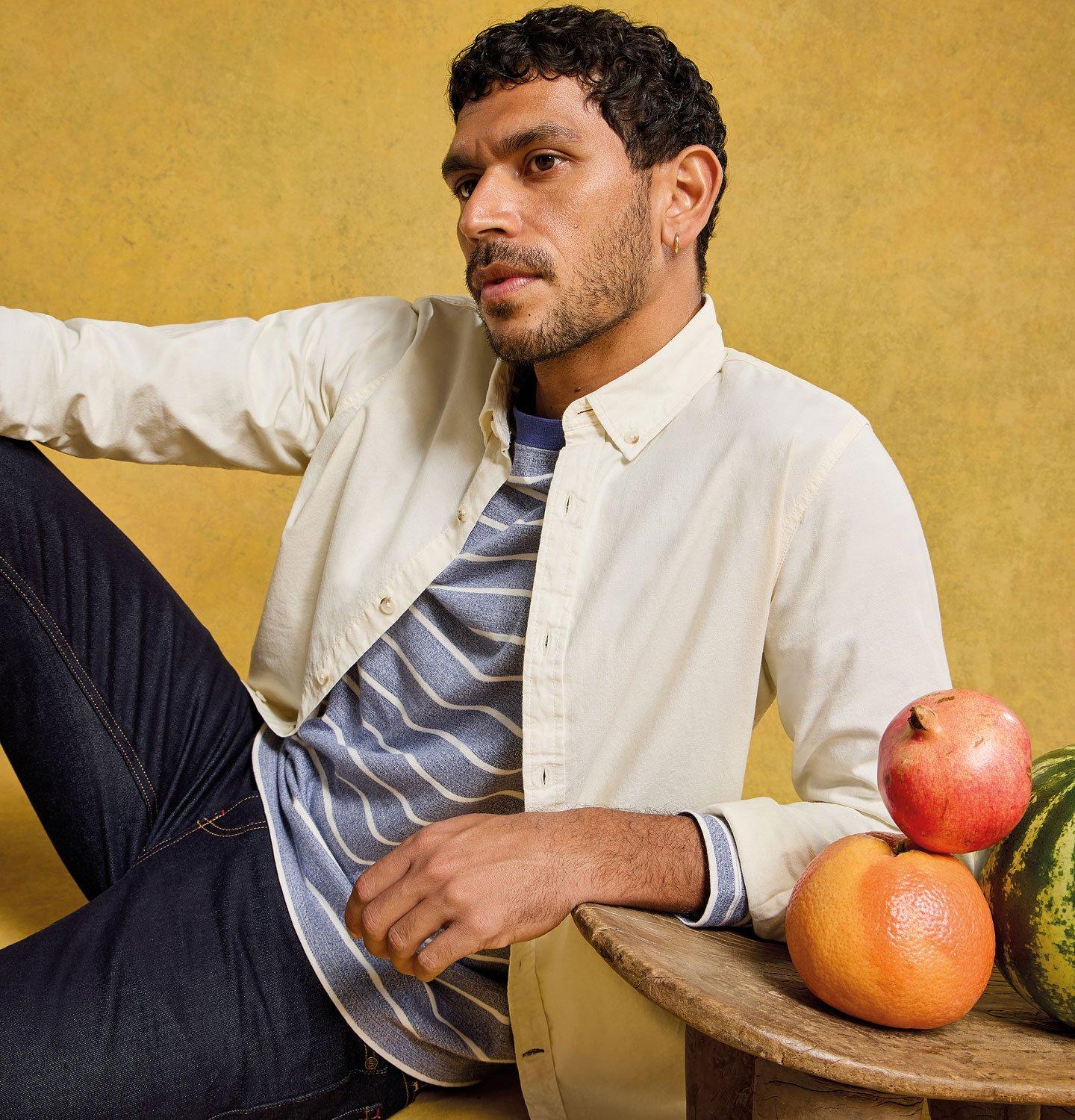a man sitting on a stool next to a table with watermelons and fruit wearing jeans, a stripey t-shirt with shirt layered on top