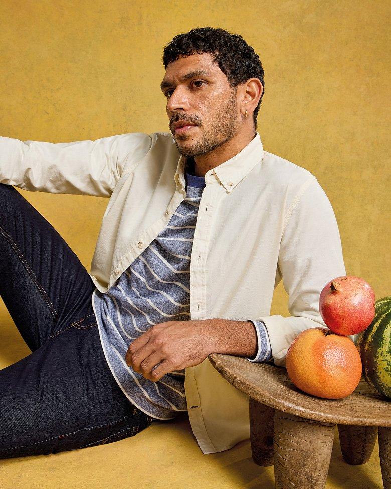 a man sitting on a stool next to a table with watermelons and fruit wearing jeans, a stripey t-shirt with shirt layered on top
