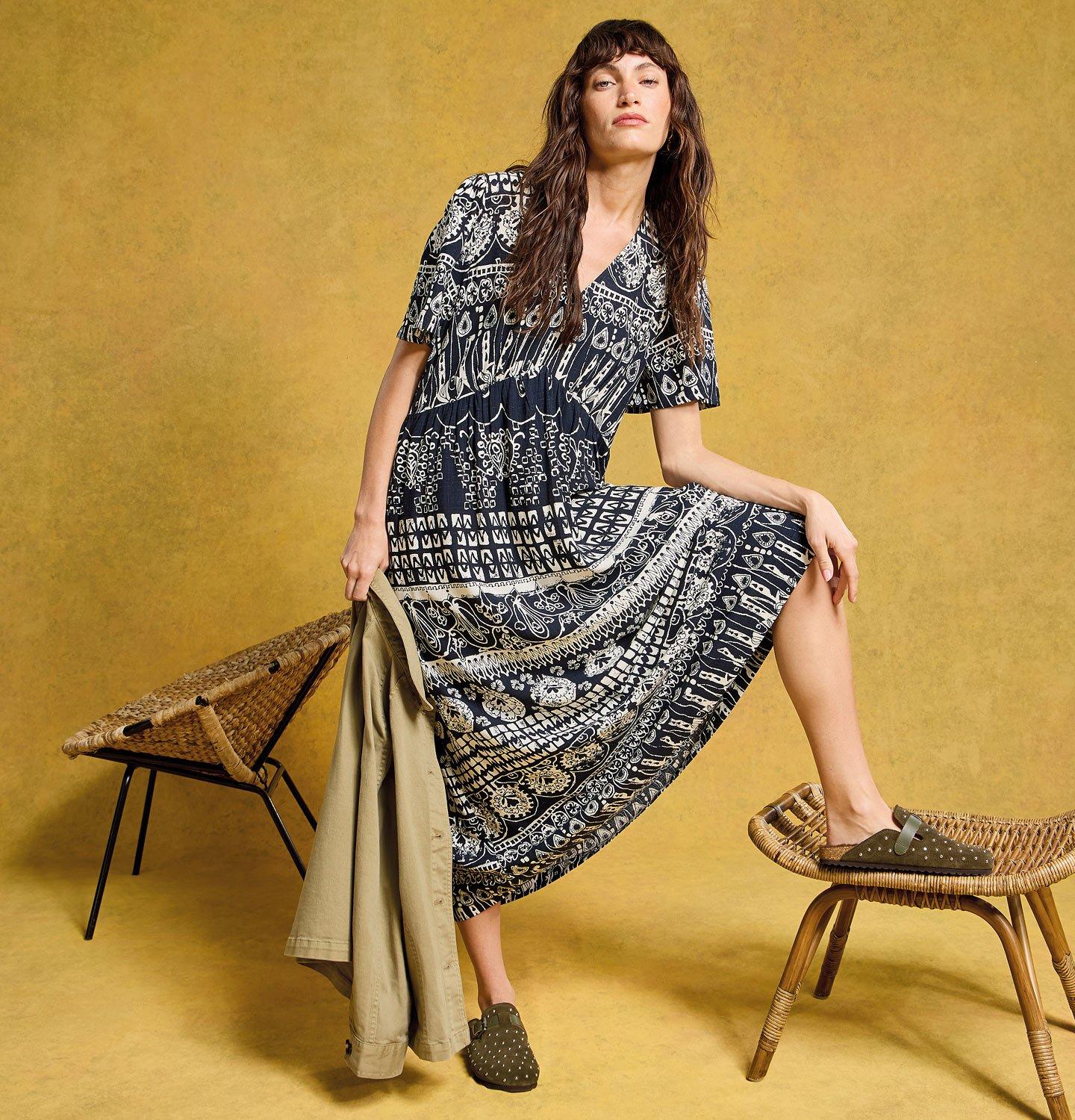 a woman in a dress holding a jacket posing on a stool