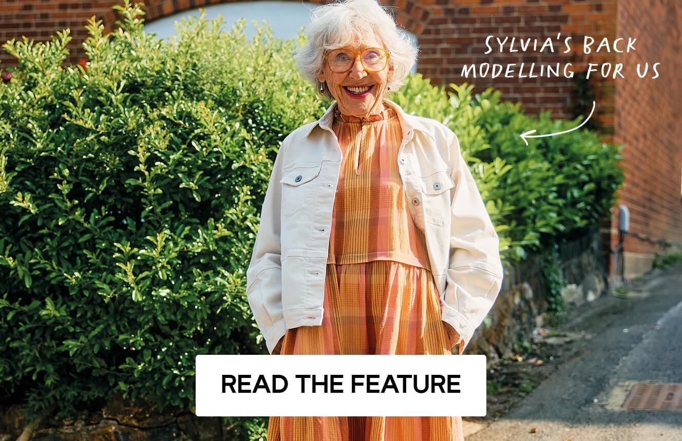 A woman wearing ab orange dress and white denim jacket standing in front of a green bush, The text says "sylvia's back modelling for us". Click here to Read the Feature.