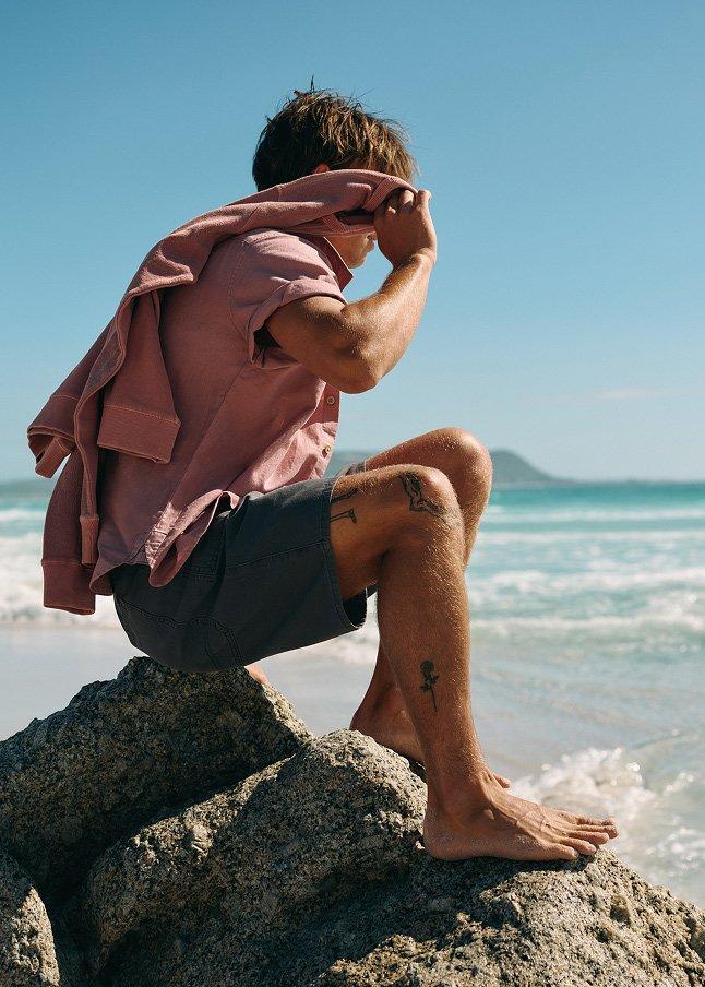 a man sitting on a rock on the beach with his shirt off