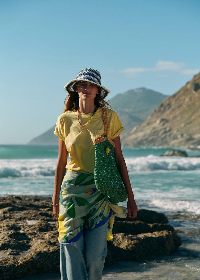 a woman walking on the beach with a hat and a bag