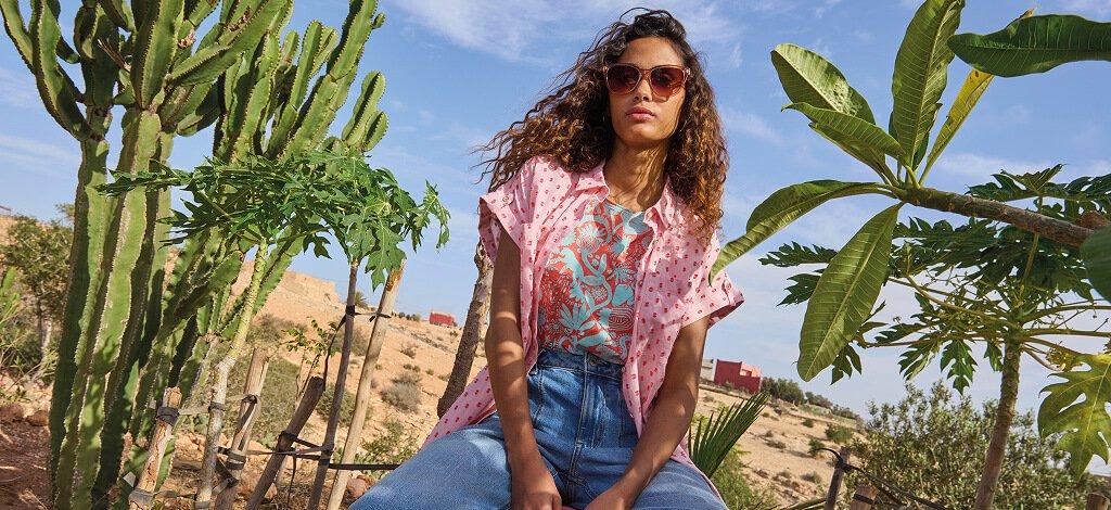 A female model sat in desert next to a cactus wearing White Stuff's ellie organic cotton shirt in pink with Tia cropped wide leg jeans. Copy reads 15% off when you sign up to emails. Click to shop.
