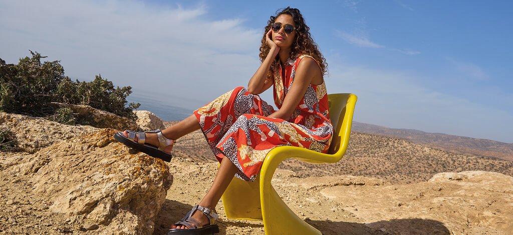 A female model sitting on a yellow chair wearing White Stuff's red Rylee Jersey Shirt Dress and pewter metallic leather sandals. Copy reads 15% off for teachers. Click to get your discount.