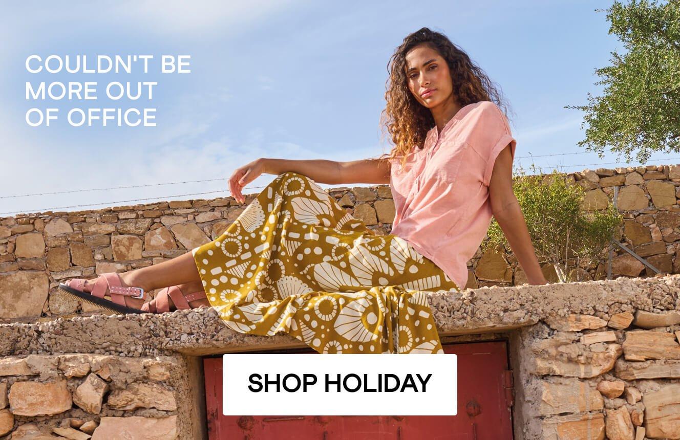 A woman sitting on a wall wearing a pink top and a patterned dress with the words 'Couldn't be more out of office' shown on the image. Click here to shop holiday clothing.