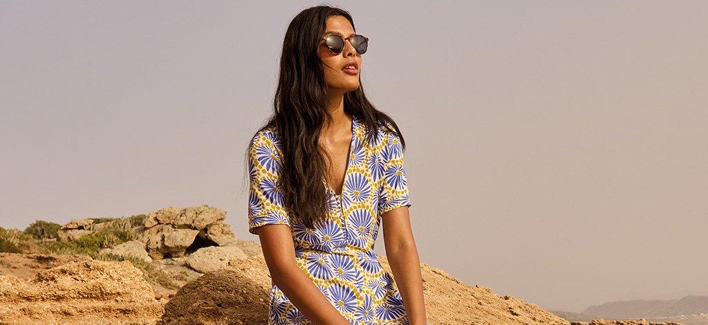 a female model standing on a box in the desert wearing White Stuff's Megan midi jersey dress in a blue print. Copy reads 15% off for NHS workers. Click to get your discount.