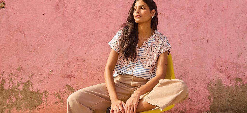 A female model sat on chair against pink wall wearing White Stuff's Nelly tee notch neck with Tia cropped wide leg jeans. Copy reads 15% off for students. Click to get your discount.