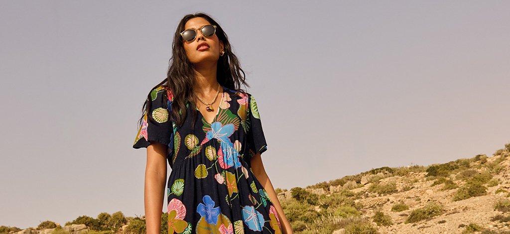 A female model stood in desert wearing White Stuff's navy multi Lucy Midi Dress. Copy reads 15% off for teachers. Click to get your discount.