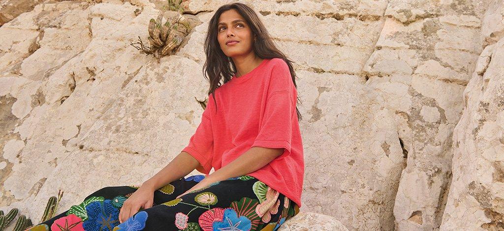 a female model sitting on a rock wearing White Stuff's Abigail navy print midi skirt styled with our Katie linen blend t-shirt in pink. Copy reads free delivery over £70. Click to shop.