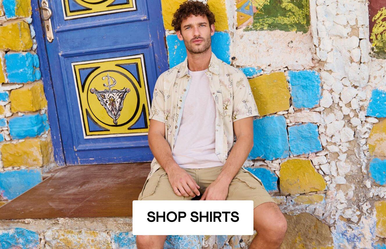 A man wearing a floral printed shirt and white tee standing in front of a colourful wall. Click here to shop men's shirts.