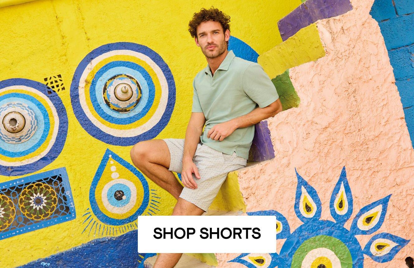 A man wearing a green polo and cream shorts sitting on steps,. Click here to shop men's shorts.
