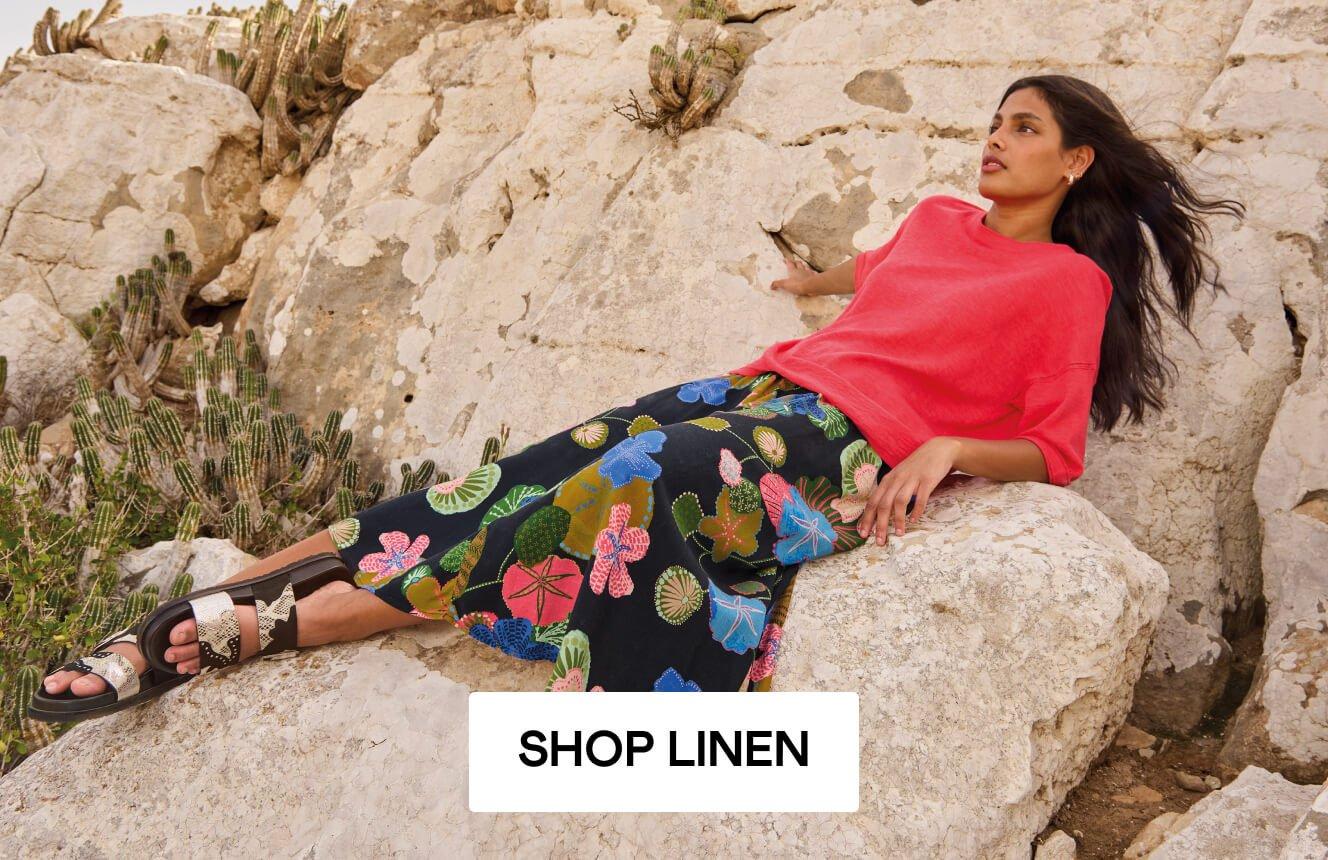 A woman wearing a pink linen top and a black skirt with geometric patterns, lounging on a rock in the desert. Click here to shop linen.