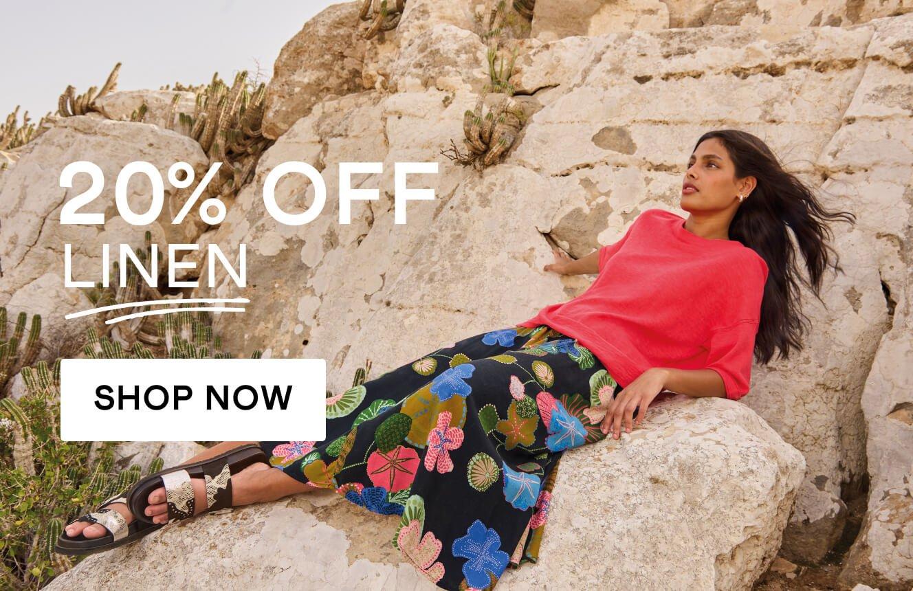 A woman wearing a pink linen top and a black skirt with geometric patterns, lounging on a rock in the desert, with the text "20% off linen", Click here to shop linen.