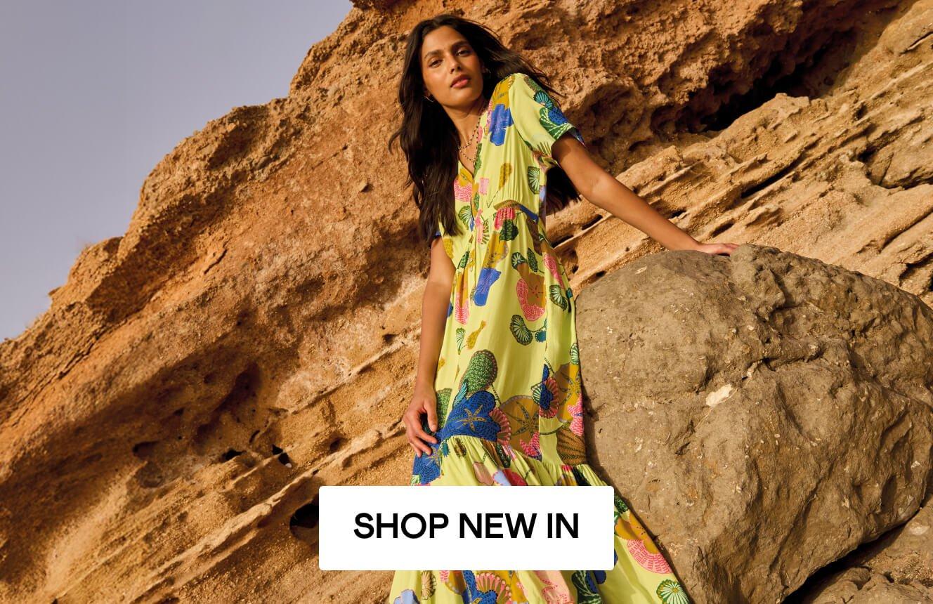 A woman wearing a lime green dress with geometric patterns, leaning on a rock in the desert, Click here to shop new in clothing. 