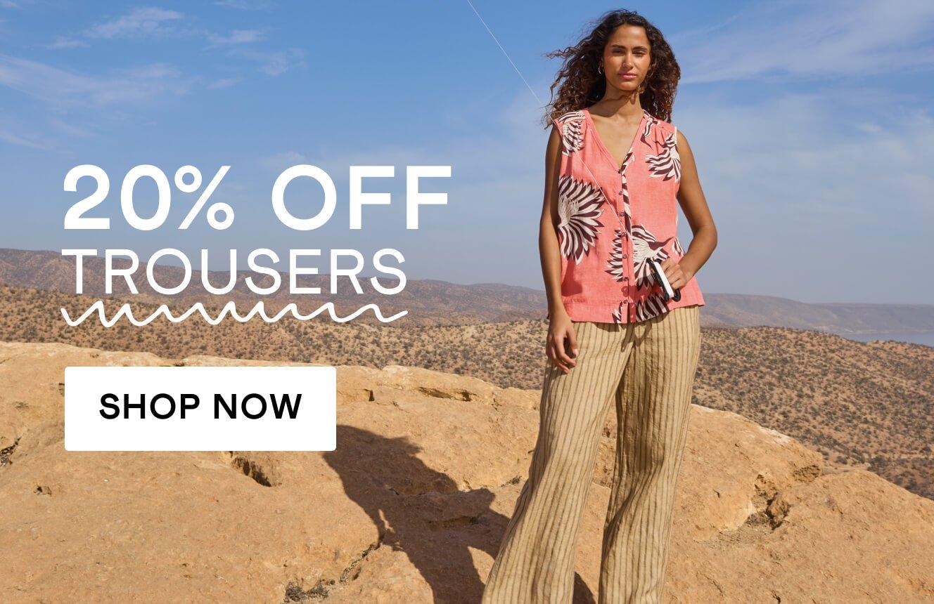 A woman wearing a coral top with a palm print and beige linen trousers with a desert background, with the text "20% off trousers", Click here to shop trousers.