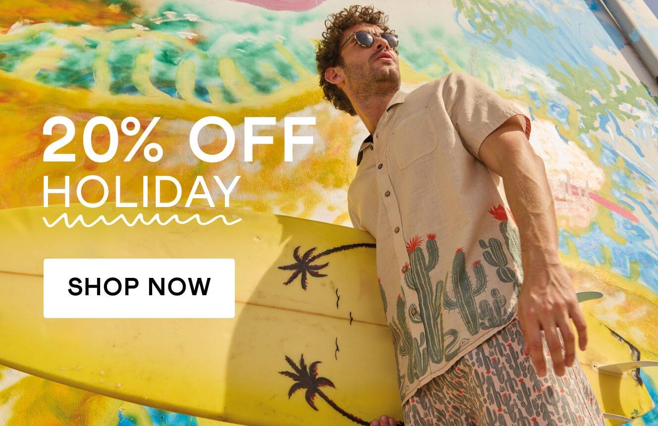 A man in a beige short sleeve shirt with a cactus print and matching shorts, holding a surfing board. Click here to shop men's holiday clothing.