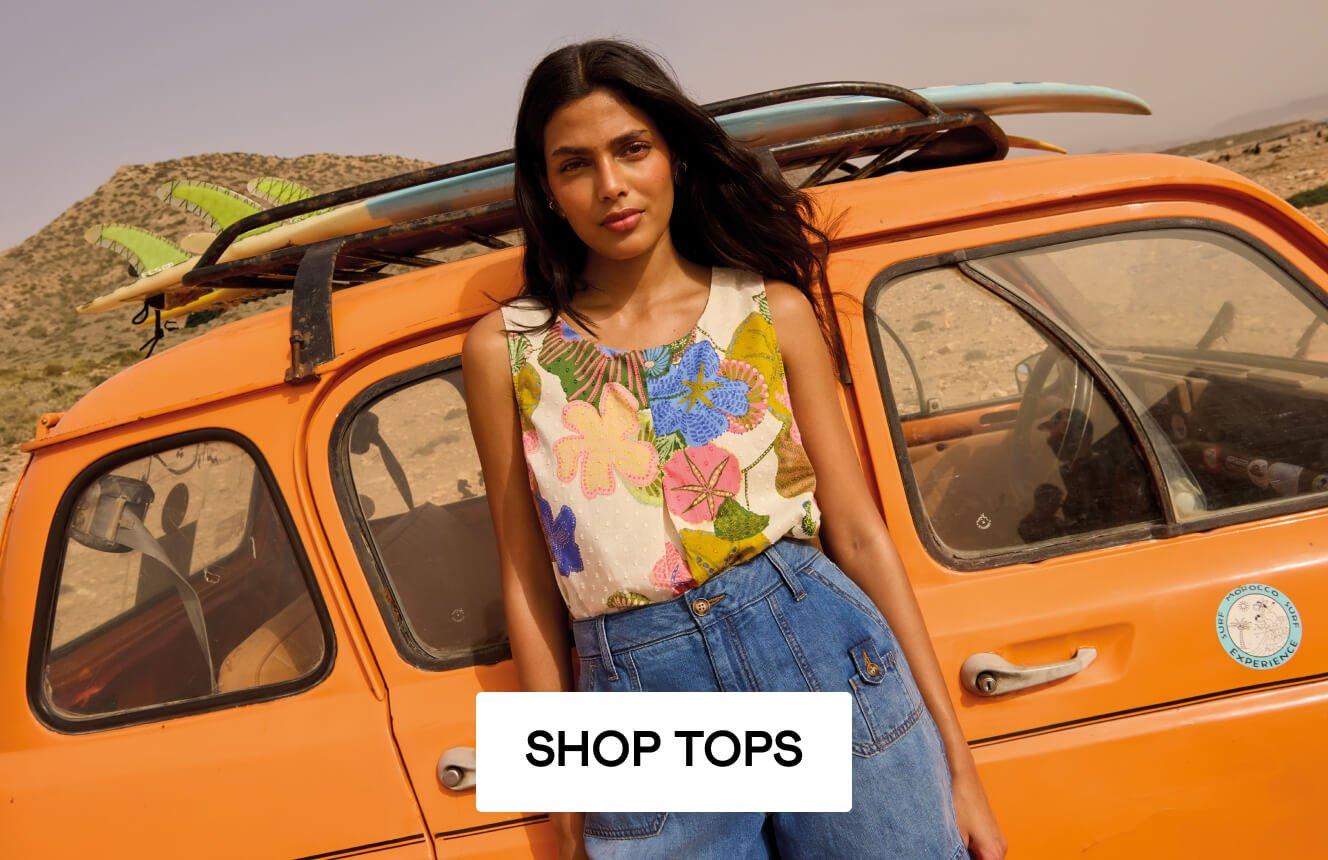 A woman wearing a floral printed top and blue denim sorts leaning on a yellow car, Click here to shop tops.