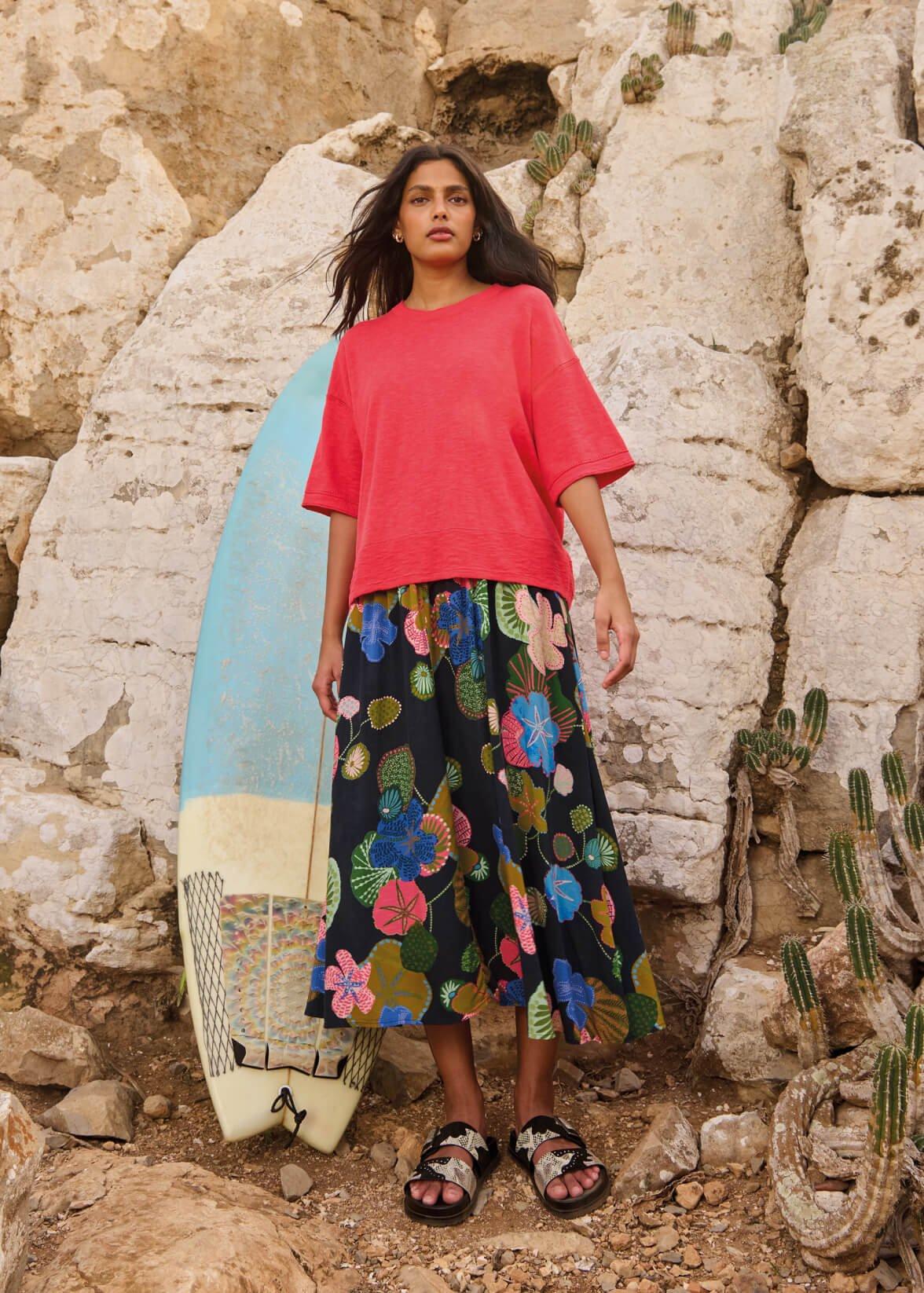 A woman wearing the kate linen blend tee and abigail print midi skirt standing infront of a surfboard