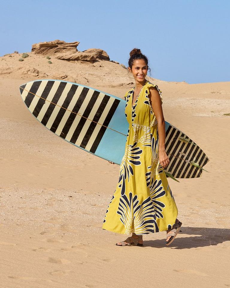 a woman walking on the beach carrying a surfboard wearing the Lorna maxi linen dress