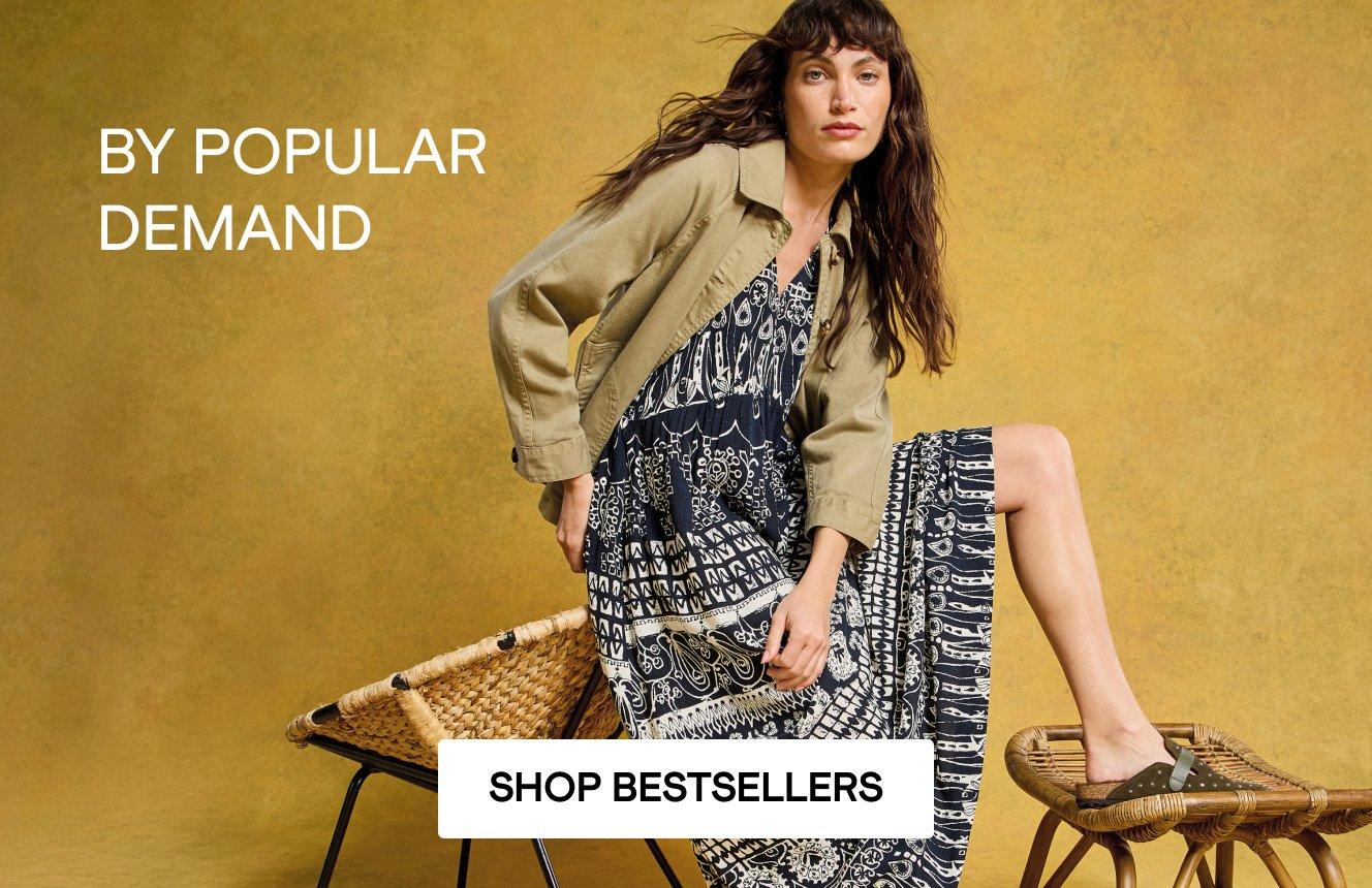 A woman wearing the black patterned Lucy Dress with the khaki Eden Jacket. Text on the image is "by popular demand". Click here to shop bestsellers.