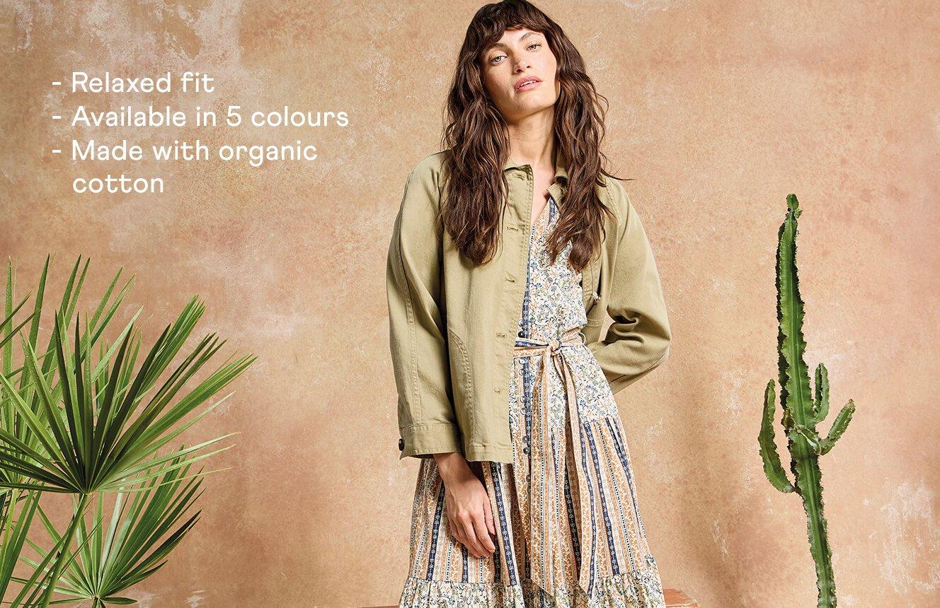 A woman wearing the green Eden Denim Jacket over the beige Clementine dress. Text on the image is "relaxed fit, available in 5 colours, made with organic cotton".
