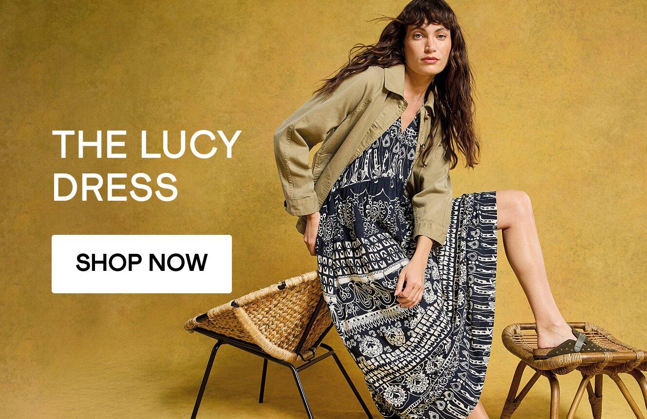 A woman wearing a black dress with geometric patterns in the desert, with the text "The Lucy Dress", Click here to shop more Lucy dresses. 