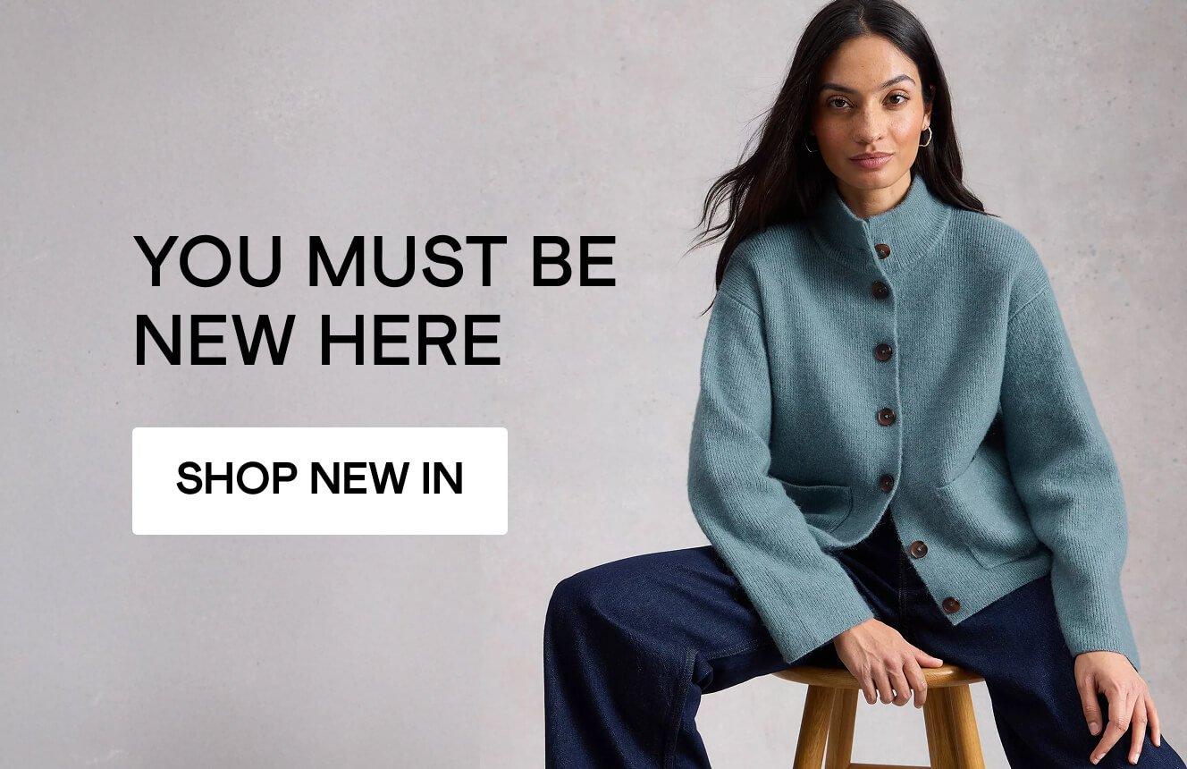 Woman wearing a teal Margot cardigan and dark pants sitting against a neutral background with the text ‘You must be new here’ and a ‘Shop New In’ button