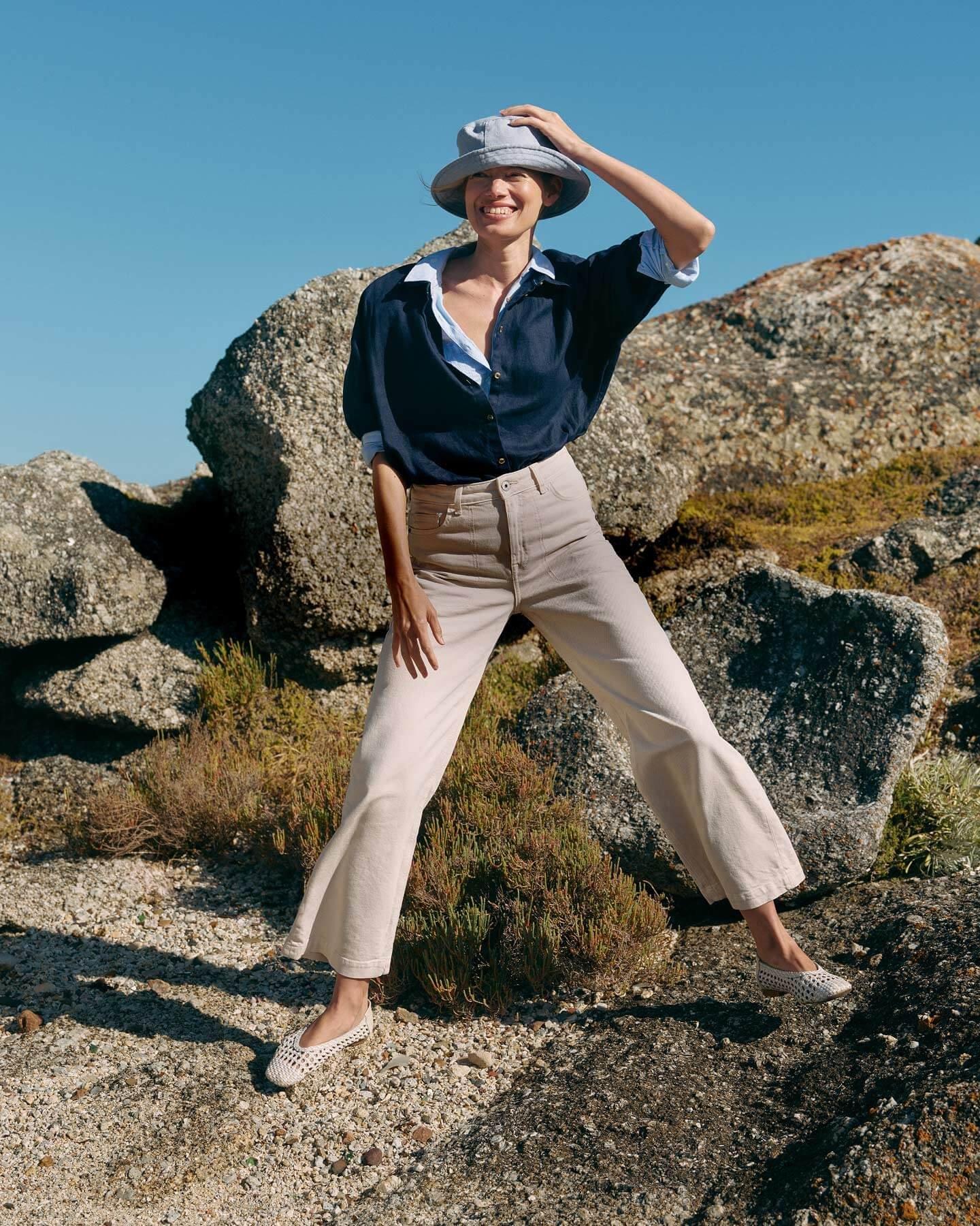 A woman stood on a rock holding a bucket hat on top of her head. She is wearing white cropped jeans, a navy blue long sleeve shirt and a pair of white woven ballerinas