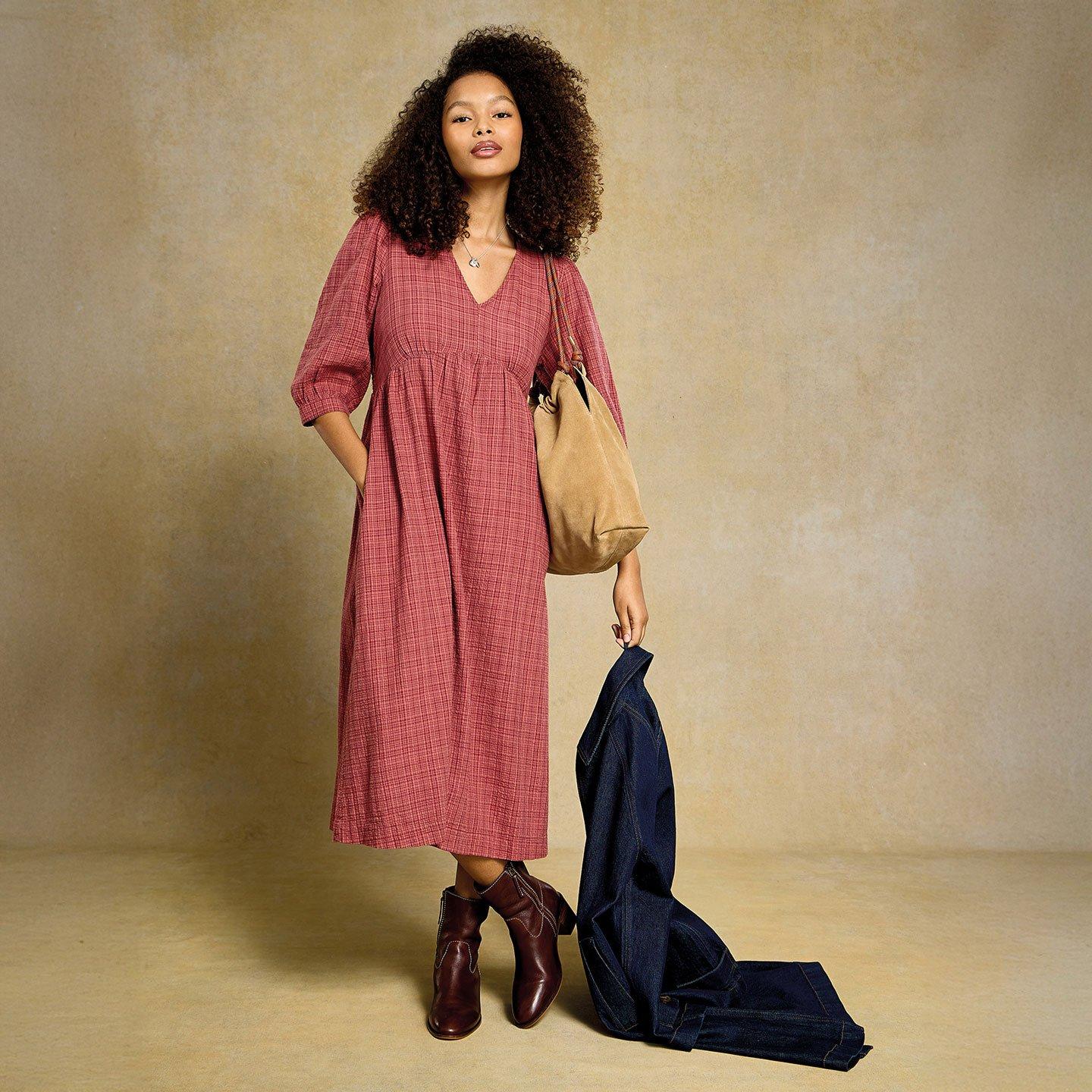 a woman with curly hair wearing a pink midi dress with a light check print, brown boots, holding a dark denim jacket posing for a photo