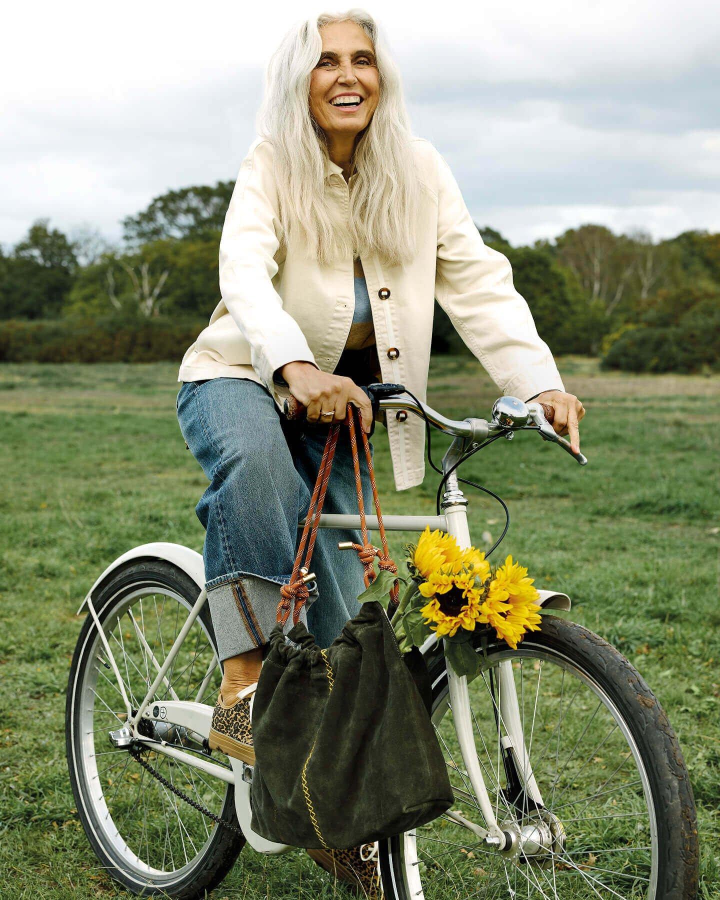 a woman posing on a bicycle outdoors wearing a pair of denim turn up jeans, a striped jumper, a cream button up jacket and leopard print trainers. A suede khaki bag is resting on the handle bars.