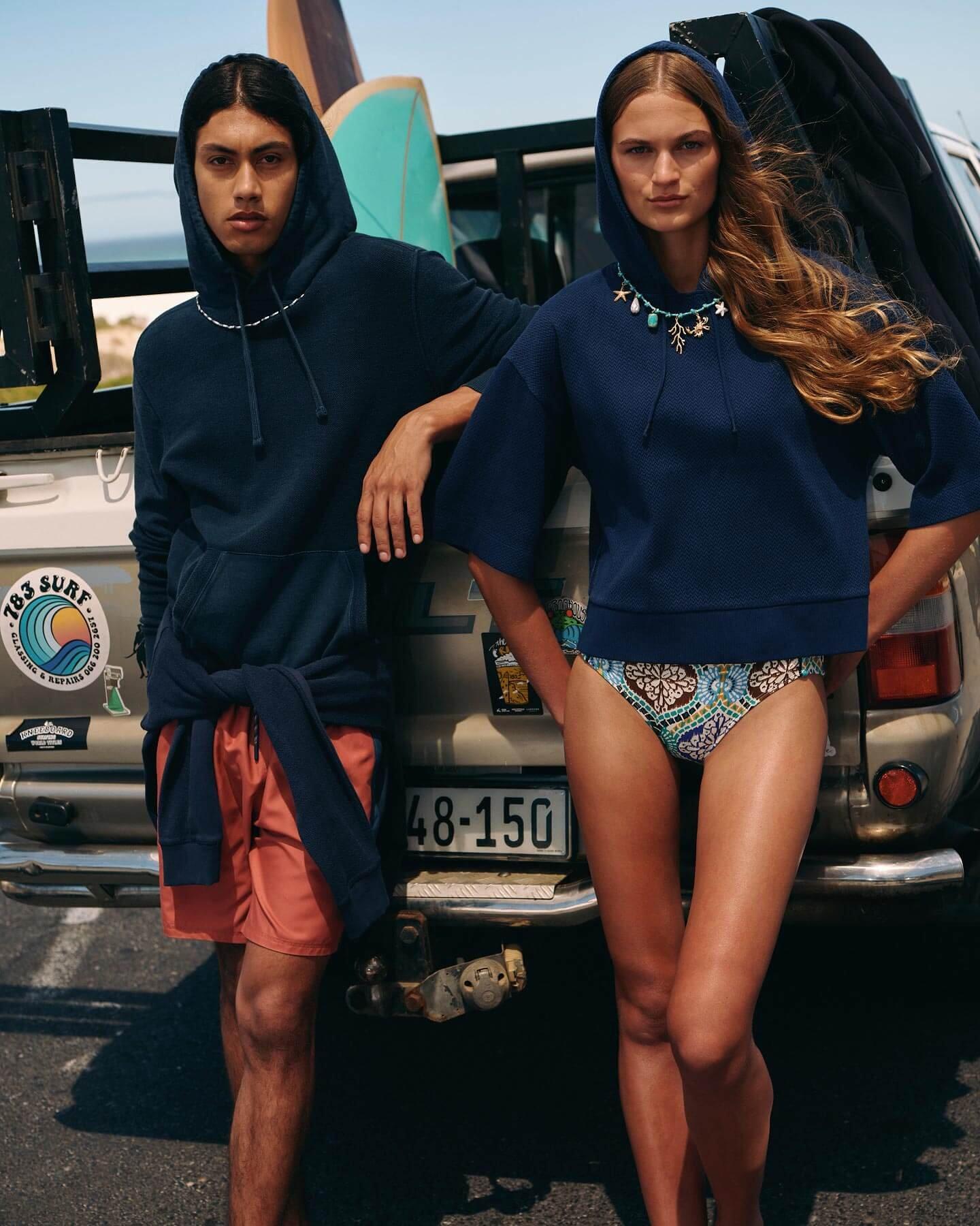 A man and woman stood against a car. She is wearing a bikini and navy short sleeve hoodie. He is wearing coral swim shorts and a navy long sleeve hoodie