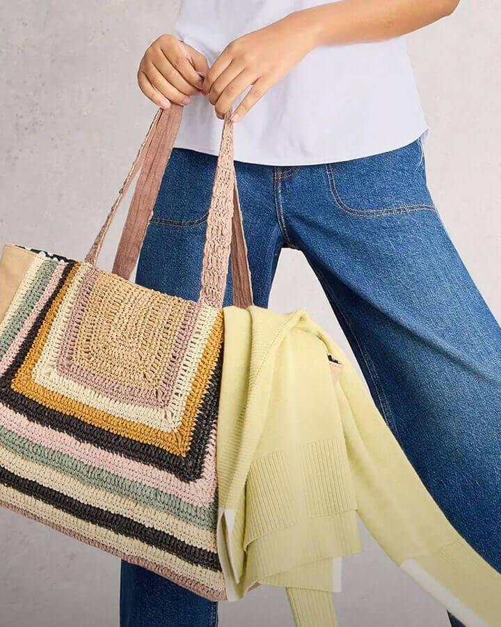 a woman stood against a grey background and is wearing denim blue jeans, a white t-shirt and is holding a colourful woven tote bag in both hands