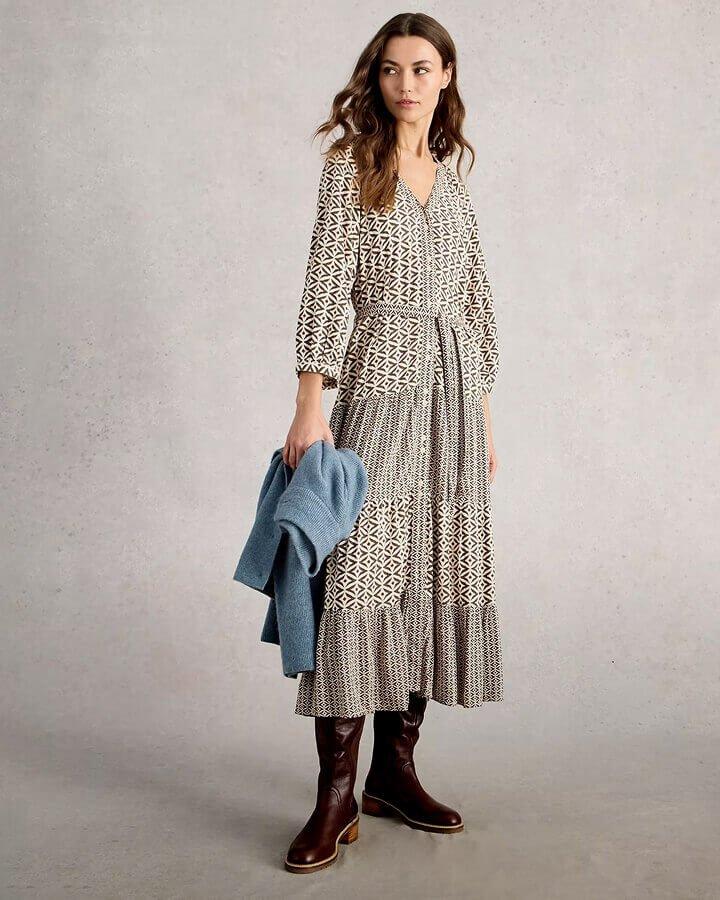 a woman stood against a grey background wearing a black printed long sleeve midi dress and a pair of brown leather mid calf boots. In her right hand she's holding a light blue cardigan.