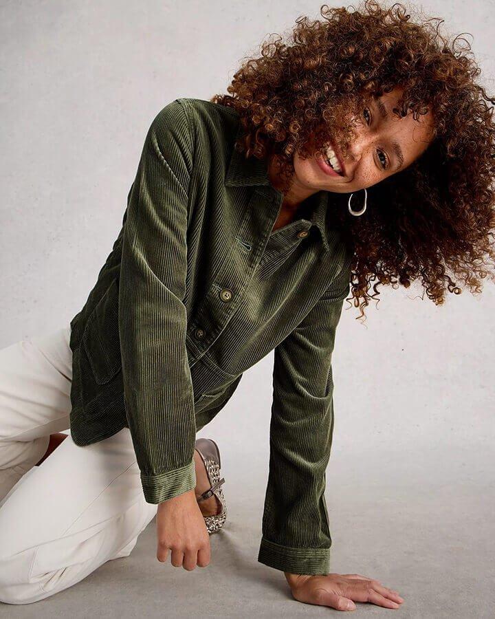 a woman kneeling down leaning on her left hand wearing a green cord jacket and a pair of white denim jeans.