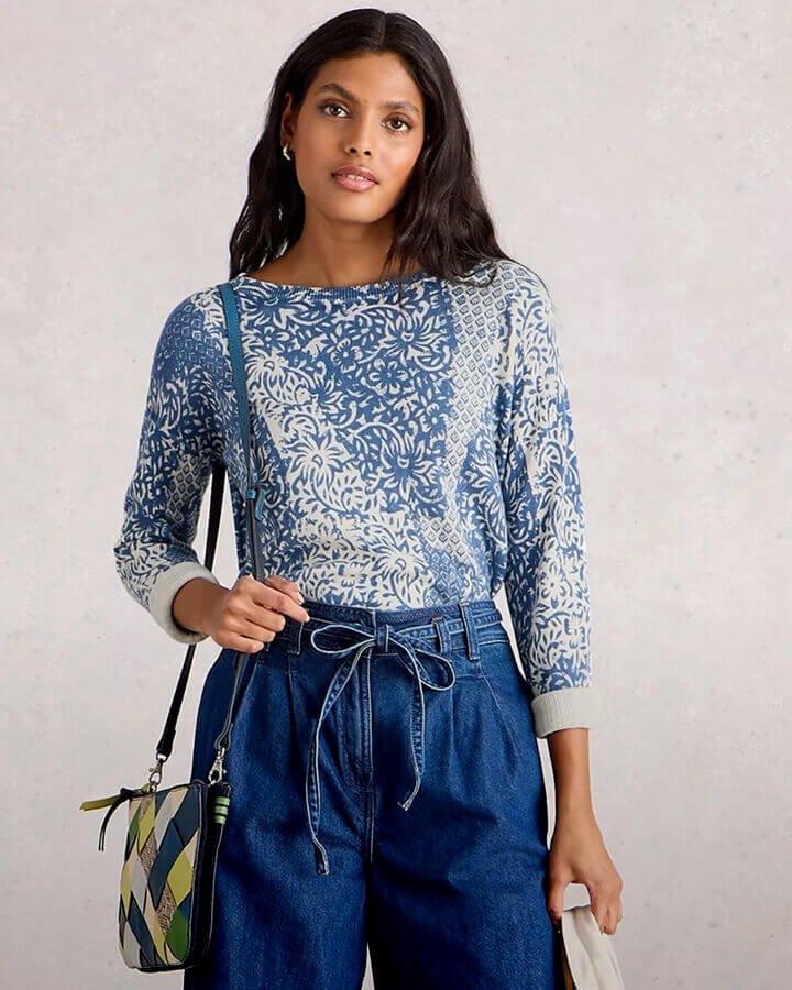 a woman stood against a grey background wearing wide leg denim jeans and a blue and white long sleeve jumper. On her right shoulder she is holding a patterned shoulder bag.