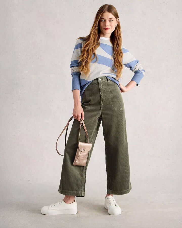 a woman stood against a grey background wearing green cord cropped trousers, a blue and white patterned jumper and a pair of white trainers. In her right hand she is holding a metallic phone bag.