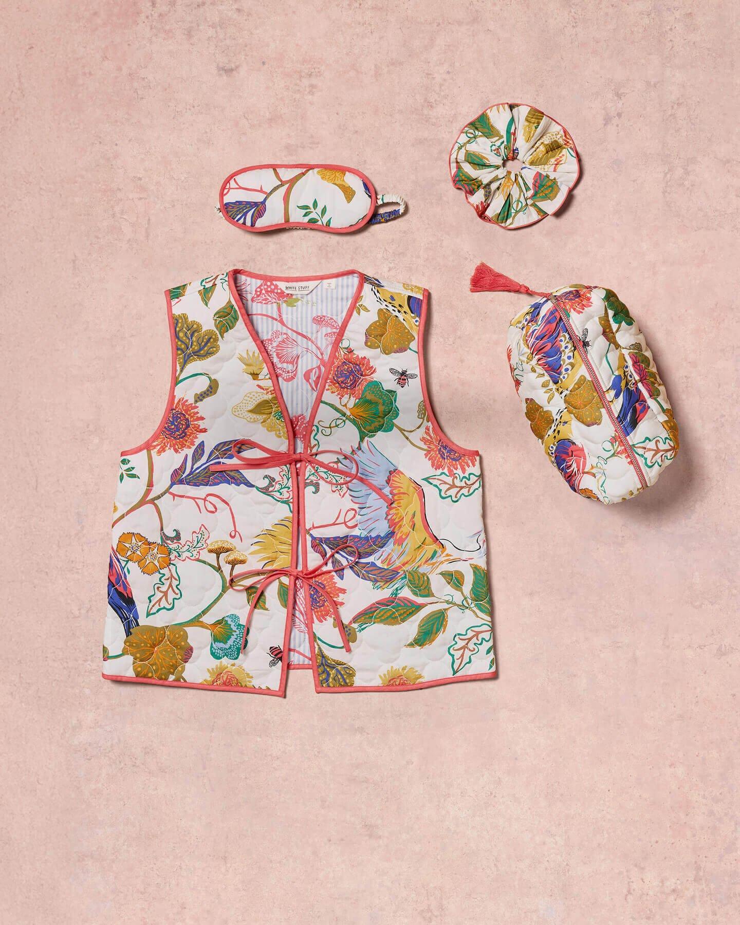 a flat lay of floral printed nightwear including a gilet, wash bag, hair scrunchie and eye mask against a pink background