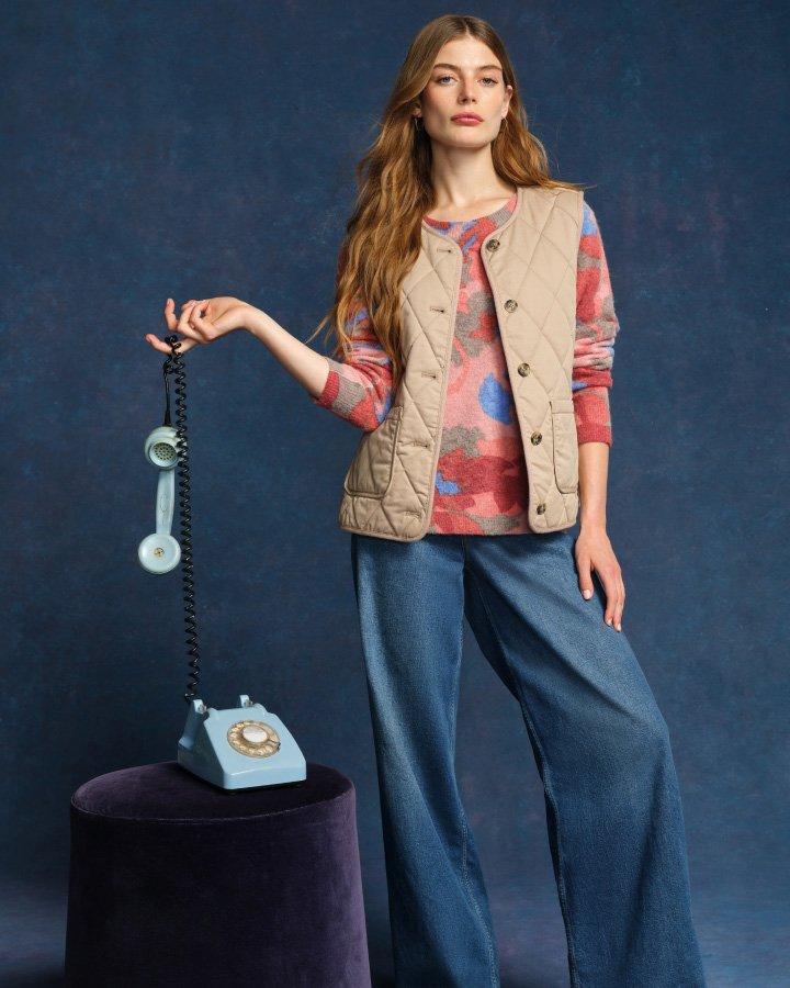 a woman standing whilst holding a telephone. Wearing a knitted jumper, jeans and a gilet